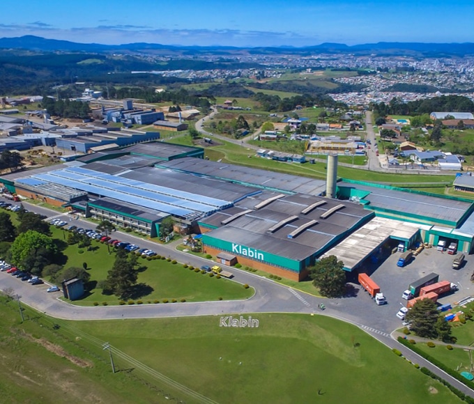 Vista aérea de uma grande unidade industrial da Klabin, com telhados verdes e cinzas, pátio com caminhões estacionados e área verde ao redor. No gramado, lê-se o nome "Klabin" em letras brancas, com uma cidade ao fundo sob céu azul.