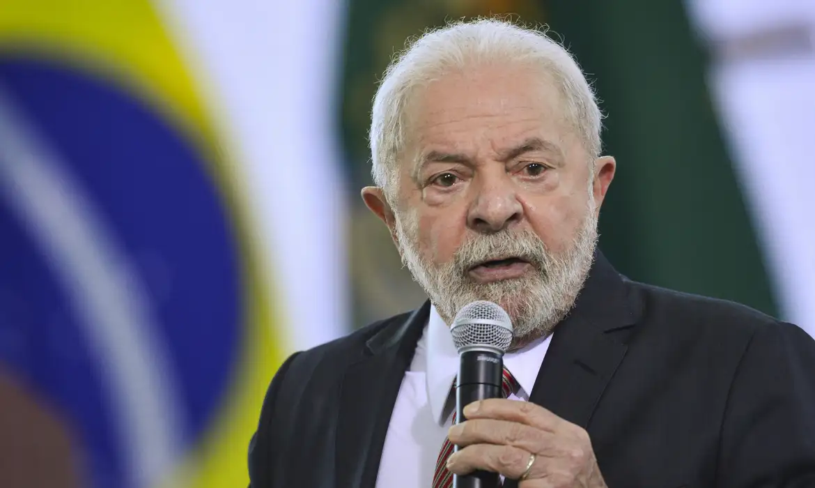 lula sancao orcamento uniao 2026 Presidente Lula discursando ao microfone, vestindo terno escuro, com a bandeira do Brasil desfocada ao fundo.