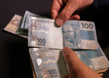 Mãos segurando diversas cédulas de 100 reais representando pagamento do INSS fevereiro 2026