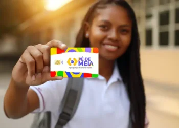 Jovem sorrindo exibe cartão colorido Pé-de-Meia na mão enquanto está na escola