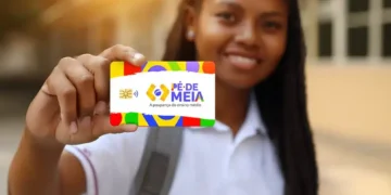 Jovem sorrindo exibe cartão colorido Pé-de-Meia na mão enquanto está na escola