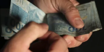 Mãos segurando e contando notas de 100 reais em fundo escuro de forma detalhada