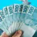 Mão segurando um leque de várias notas de 100 reais sobre um fundo azul vibrante, representando o pagamento do benefício.
