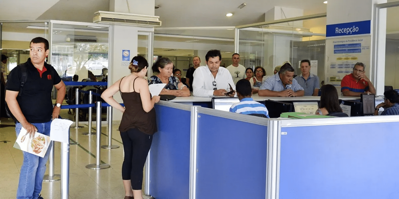 Pessoas aguardando atendimento em agência do INSS com guichês azuis e placa de recepção ao fundo.