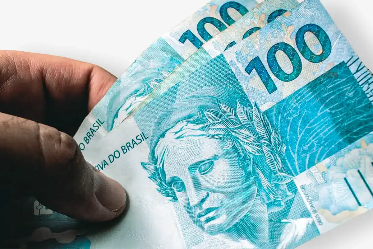 Valores Bolsa Família 2026 Mão segurando três notas de 100 reais representando os valores do Bolsa Família em 2026