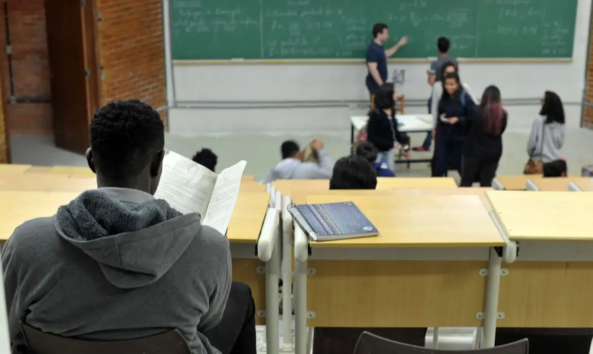 Aula com estudante lendo material Estudante sentado em uma sala de aula lendo um papel, com professor escrevendo no quadro negro à frente