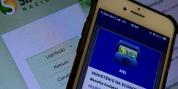 Tela de smartphone exibindo o logotipo azul do aplicativo MEI da Receita Federal, posicionado à frente de um monitor com o site do Simples Nacional ao fundo.