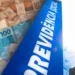 Composição visual com notas de 50 e 100 reais espalhadas ao fundo e uma placa azul vertical em primeiro plano com a palavra "Previdência" escrita em letras brancas.