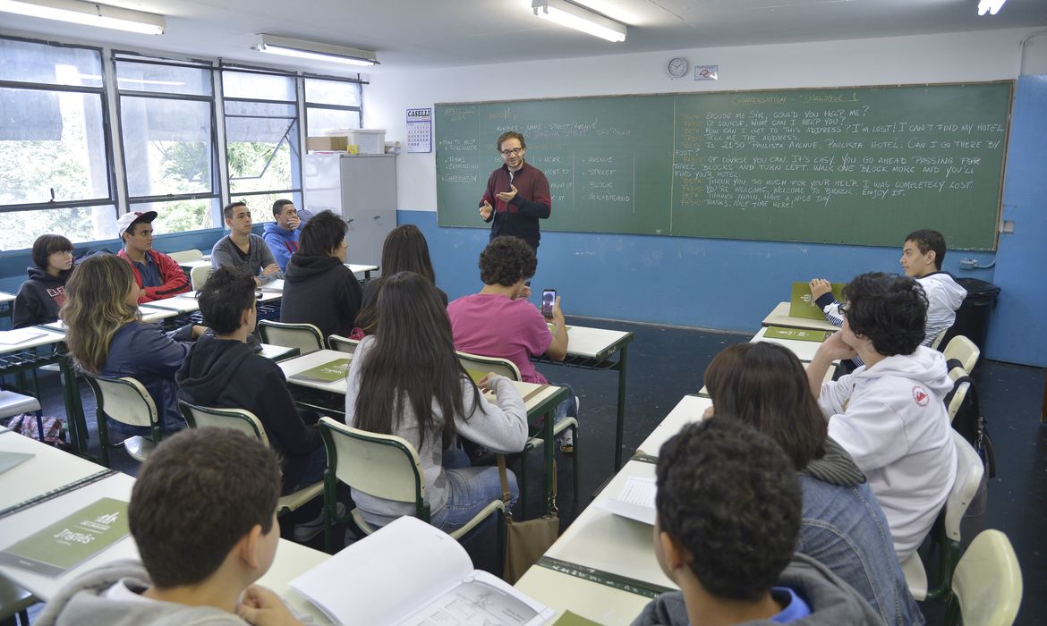 Professor em sala de aula gesticulando enquanto explica o conteúdo para alunos sentados em fileiras, ilustrando a rotina beneficiada pela nova Medida Provisória.