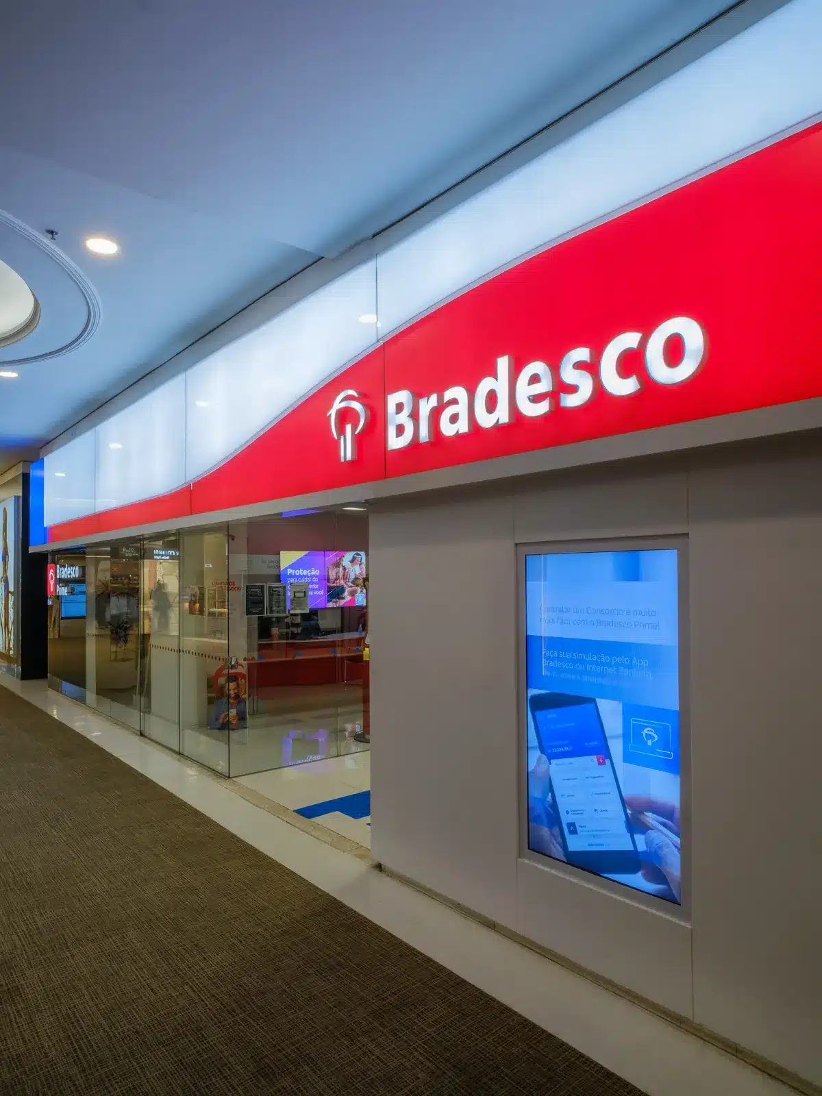 Agência Bradesco com vagas abertas. Fachada de agência Bradesco com logo iluminado e tela digital com informações do banco.