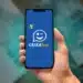 Mão segurando um smartphone que exibe o logotipo do aplicativo CAIXA Tem (um ícone de chat piscando) em uma tela azul, com notas de 100 reais borradas ao fundo.