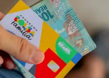 Cartão do Bolsa Família junto a cédula de dinheiro ilustrando renda familiar dentro do limite do programa.