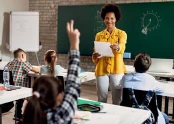 Fotografia de uma professora negra sorridente vestindo blusa amarela em uma sala de aula. Ela segura um papel e gesticula para os alunos sentados. Em primeiro plano, uma criança levanta a mão para participar da aula.