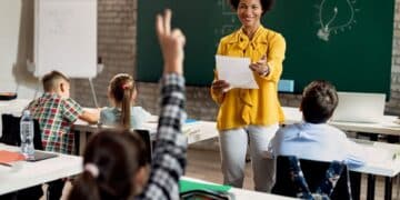 Fotografia de uma professora negra sorridente vestindo blusa amarela em uma sala de aula. Ela segura um papel e gesticula para os alunos sentados. Em primeiro plano, uma criança levanta a mão para participar da aula.