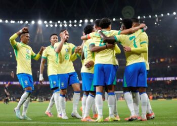 Jogadores da Seleção Brasileira de futebol, vestindo o uniforme oficial amarelo e azul, abraçados em comemoração no centro de um gramado de estádio iluminado à noite.