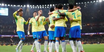 Jogadores da Seleção Brasileira de futebol, vestindo o uniforme oficial amarelo e azul, abraçados em comemoração no centro de um gramado de estádio iluminado à noite.