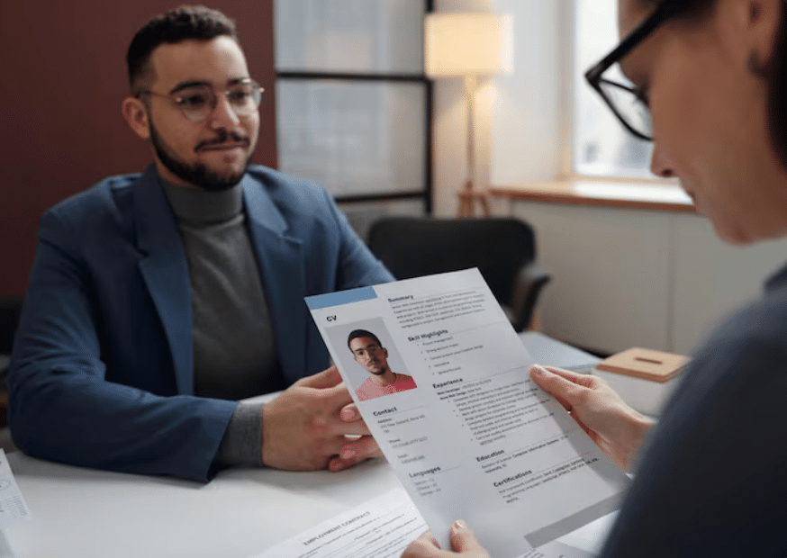 Uma recrutadora segura um currículo impresso com a foto de um candidato jovem de óculos. Ao fundo, o candidato observa com um leve sorriso enquanto o documento, que contém seções de "Resumo" e "Habilidades", é analisado.