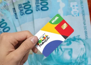 Mão de uma pessoa segurando o cartão oficial do Programa Bolsa Família com chip, posicionado sobre várias notas de 100 reais espalhadas ao fundo.