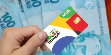 Mão de uma pessoa segurando o cartão oficial do Programa Bolsa Família com chip, posicionado sobre várias notas de 100 reais espalhadas ao fundo.