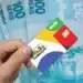 Mão de uma pessoa segurando o cartão oficial do Programa Bolsa Família com chip, posicionado sobre várias notas de 100 reais espalhadas ao fundo.