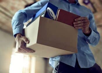 Homem vestindo camisa azul segurando uma caixa de papelão com pertences de escritório, como pastas e cadernos, em um ambiente interno iluminado.