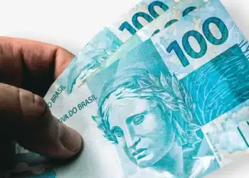 Notas de R$ 100 sendo seguradas por uma mão, representando recursos esquecidos em bancos.