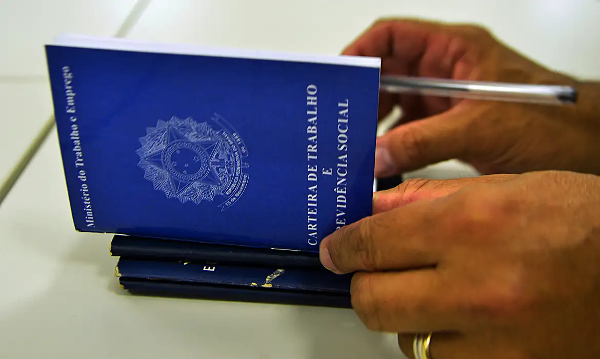 guia atualizado clt direitos trabalhistas 2026 Close-up de mãos masculinas manuseando uma Carteira de Trabalho e Previdência Social azul aberta; ao fundo, uma caneta repousa sobre a mesa de atendimento.