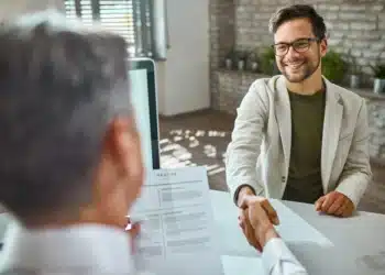 Homem em entrevista de emprego apertando as mãos com o entrevistador, enquanto apresenta seu currículo.