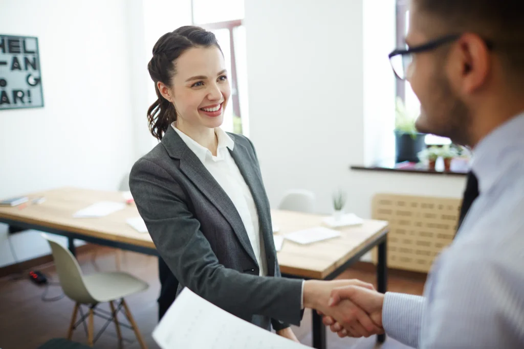 Dicas para conquistar uma vaga de emprego em 2026 Mulher apertando as mãos durante uma entrevista de emprego, sorrindo e se apresentando.