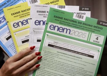 Mão segurando diversos cadernos de prova do Enem 2025 nas cores azul, amarelo, branco e verde, com foco no caderno verde do primeiro dia.