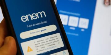 Mão segurando um celular que exibe a tela de login do aplicativo do Enem, com o botão "Entrar com gov.br" em destaque e uma página web ao fundo.