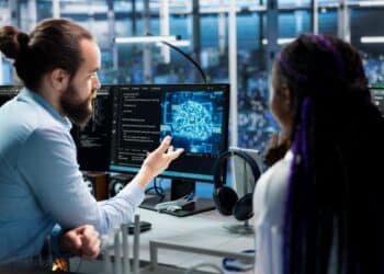 Um homem com cabelo preso em um coque e barba aponta para um monitor que exibe a representação gráfica de um cérebro digital azul com a sigla "AI". À direita, uma mulher com tranças roxas observa a explicação em um escritório de tecnologia moderno.