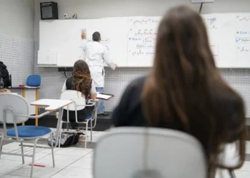 Professor escrevendo fórmulas químicas no quadro branco em sala com estudantes atentos sentados