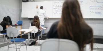 Professor escrevendo fórmulas químicas no quadro branco em sala com estudantes atentos sentados