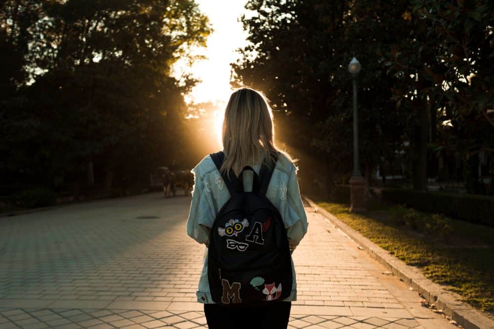 Vista traseira de uma estudante caminhando por uma calçada ensolarada, usando jaqueta jeans e mochila preta com adesivos, simbolizando a evasão escolar.
