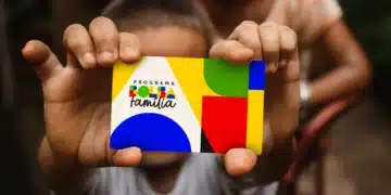 Close das mãos de uma criança segurando o cartão colorido do Programa Bolsa Família em destaque, com o fundo desfocado mostrando uma pessoa e um ambiente doméstico.