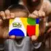 Close das mãos de uma criança segurando o cartão colorido do Programa Bolsa Família em destaque, com o fundo desfocado mostrando uma pessoa e um ambiente doméstico.