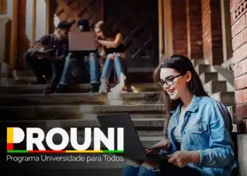 Estudante utilizando um laptop enquanto outros colegas estão atrás dela, em um ambiente acadêmico com escadas e paredes de tijolos. A imagem representa um momento de estudo em grupo.