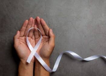 Duas mãos abertas segurando um laço branco, símbolo da campanha Janeiro Branco, sobre um fundo cinza de textura rústica.