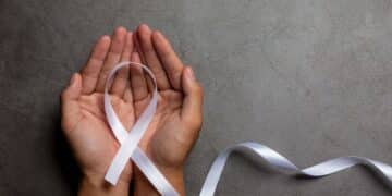 Duas mãos abertas segurando um laço branco, símbolo da campanha Janeiro Branco, sobre um fundo cinza de textura rústica.