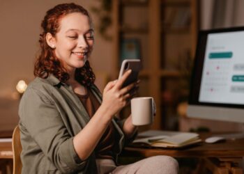 Mulher jovem com cabelos ruivos sorrindo ao olhar para o celular enquanto segura uma caneca; ao fundo, um monitor de computador exibe um calendário digital organizado.