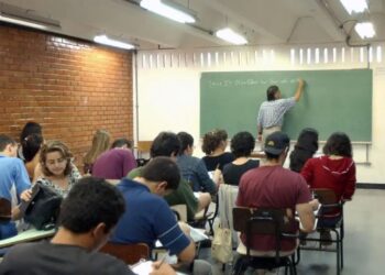 Texto Alternativo (Alt Text): Professor de costas escrevendo no quadro negro em uma sala de aula com alunos adultos sentados em suas carteiras, simbolizando a educação básica.