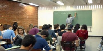 Texto Alternativo (Alt Text): Professor de costas escrevendo no quadro negro em uma sala de aula com alunos adultos sentados em suas carteiras, simbolizando a educação básica.