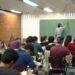 Texto Alternativo (Alt Text): Professor de costas escrevendo no quadro negro em uma sala de aula com alunos adultos sentados em suas carteiras, simbolizando a educação básica.