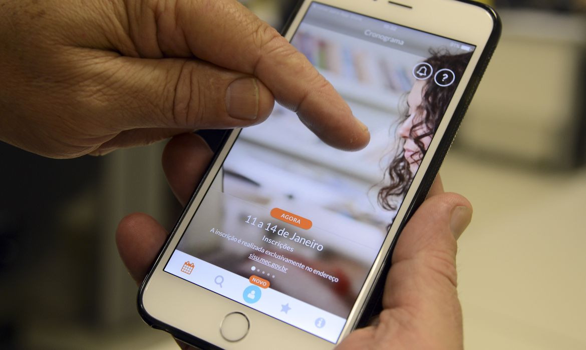 Close de uma mão masculina segurando um iPhone branco, exibindo o aplicativo oficial do MEC com o cronograma das inscrições do SiSU na tela.
