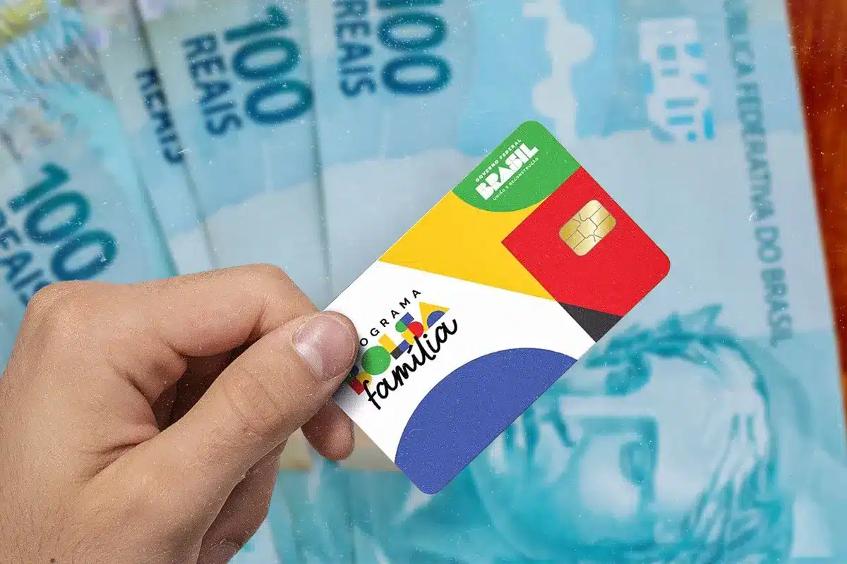quem tem direito pagamento parcial bolsa familia 2026. Mão masculina segurando o novo cartão magnético do Programa Bolsa Família, com design colorido em amarelo, azul e vermelho, posicionado à frente de várias cédulas de 100 reais ao fundo.