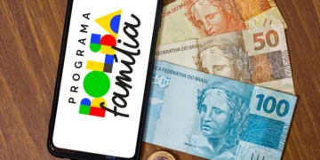 Smartphone sobre uma mesa de madeira exibindo o logotipo colorido do Programa Bolsa Família na tela, posicionado ao lado de cédulas de 100, 50 e 20 reais e algumas moedas.