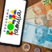 Smartphone sobre uma mesa de madeira exibindo o logotipo colorido do Programa Bolsa Família na tela, posicionado ao lado de cédulas de 100, 50 e 20 reais e algumas moedas.