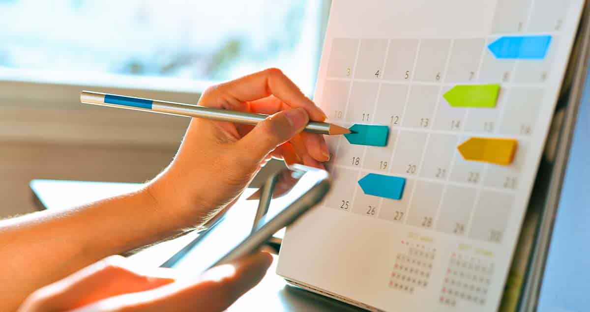 Pessoa marcando no calendário com lápis colorido e segurando um celular, simbolizando a organização e planejamento de metas.