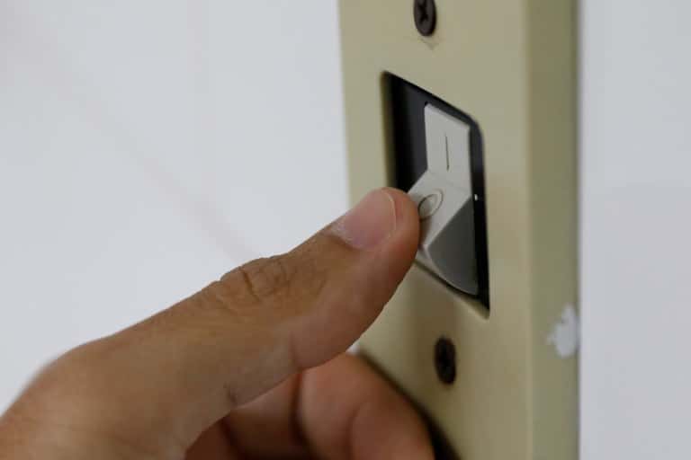 Close do dedo de uma pessoa pressionando um interruptor de luz branco na parede para desligar ou ligar a energia. O foco está no contato do dedo com a tecla do interruptor, simbolizando o controle do consumo elétrico.